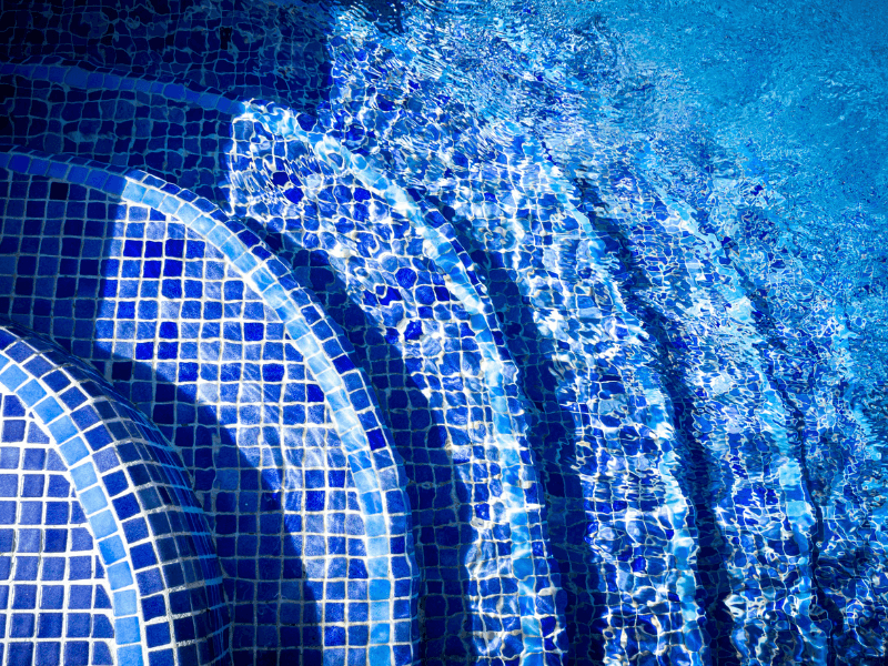Close-up of submerged curved pool steps covered in blue and white mosaic tiles with rippling water above.